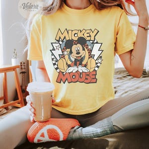 Puede incluir: Camiseta amarilla con un gr&aacute;fico de Mickey Mouse y el texto "Mickey Mouse". El dise&ntilde;o tiene un estilo retro con estrellas, rayos y un patr&oacute;n de cuadros. La camiseta est&aacute; hecha de un material suave y c&oacute;modo.