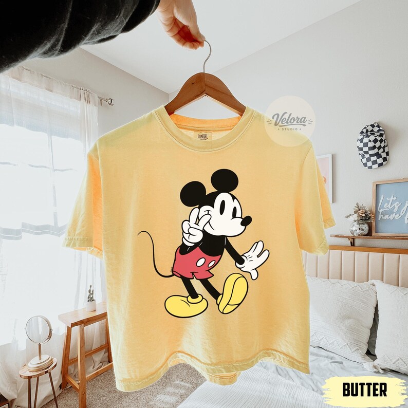 May include: A yellow t-shirt featuring a graphic of Mickey Mouse in a classic pose. The cartoon character is depicted in black, white, red, and yellow. The word "BUTTER" is printed in a yellow rectangle at the bottom of the shirt.