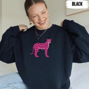 May include: Black sweatshirt featuring a pink leopard design. The leopard has dark pink spots and a light pink body. The word "BLACK" is printed in white in the upper right corner. The sweatshirt is a casual style.