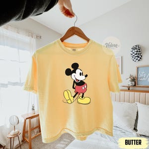 May include: A yellow t-shirt with a classic Mickey Mouse graphic. The cartoon character is depicted in black, red, and yellow. The shirt is hanging on a wooden hanger. The word "BUTTER" is printed on the bottom of the image.