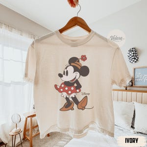 May include: Ivory-colored t-shirt featuring a classic Minnie Mouse graphic. The design shows Minnie in a red polka-dot skirt, brown shoes, and a matching hat. The word "Disney" is printed below the character.
