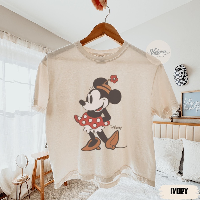 May include: Ivory-colored t-shirt featuring a classic Minnie Mouse graphic. The design shows Minnie in a red polka-dot skirt, brown shoes, and a matching hat. The word "Disney" is printed below the character.