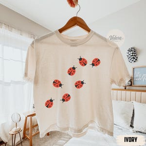 May include: An ivory short-sleeved t-shirt featuring a scattered pattern of small red ladybugs with black spots. The shirt has a crew neckline and is displayed on a wooden hanger against a light-colored interior setting with soft lighting.