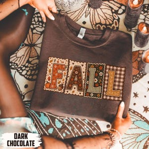 May include: A dark chocolate brown crewneck sweatshirt featuring the word FALL in large block letters. Each letter has a unique pattern including floral prints, autumn leaves, pumpkins, acorns, and a brown and white checkered design.
