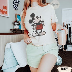 Może przedstawiać: Kremowa koszulka z nadrukiem Mickey Mouse w czerwonych spodenkach. Koszulka jest połączona z jasnozielonymi szortami. Zdjęcie zrobione w pomieszczeniu, z dekoracyjną ścianą i kulą dyskotekową w tle.