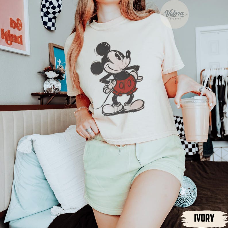 Może przedstawiać: Kremowa koszulka z nadrukiem Mickey Mouse w czerwonych spodenkach. Koszulka jest połączona z jasnozielonymi szortami. Zdjęcie zrobione w pomieszczeniu, z dekoracyjną ścianą i kulą dyskotekową w tle.