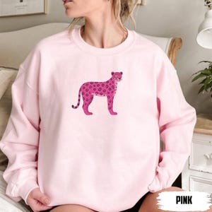 May include: A light pink sweatshirt featuring a pink leopard illustration. The leopard has dark pink spots and a black striped tail. The word "PINK" is printed in a bold, white font at the bottom right corner of the image.