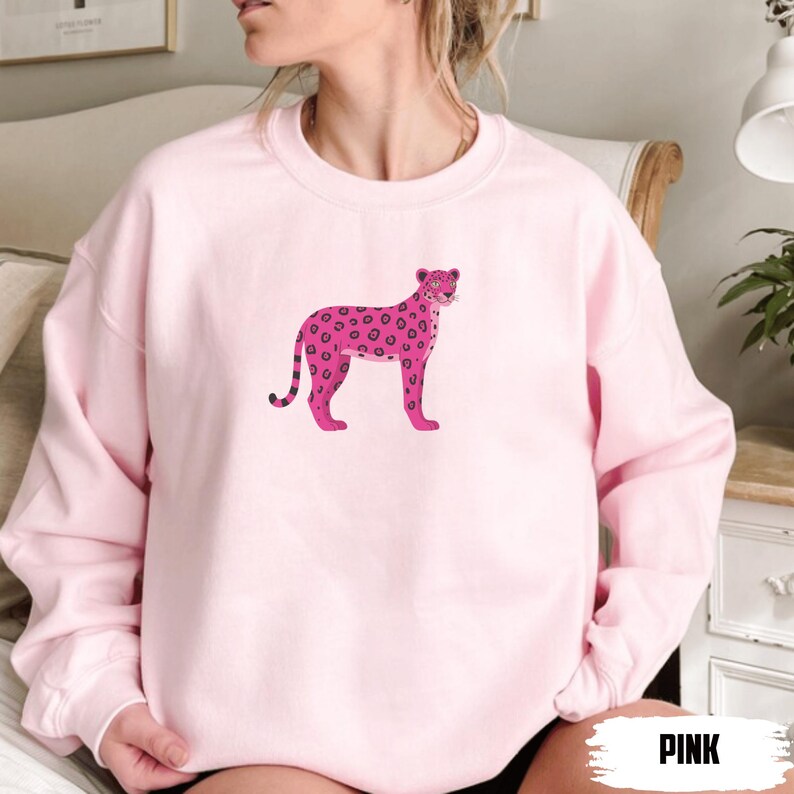 May include: A light pink sweatshirt featuring a pink leopard illustration. The leopard has dark pink spots and a black striped tail. The word "PINK" is printed in a bold, white font at the bottom right corner of the image.