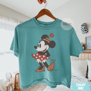 May include: A teal-colored t-shirt featuring a cartoon illustration of Minnie Mouse. Minnie is depicted in a classic pose, wearing a red polka-dot skirt, brown shoes, and a hat with a flower. The word "Disney" is printed below the character.