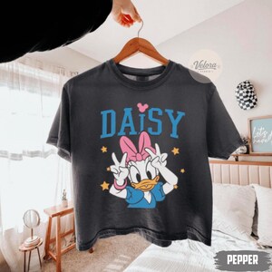 May include: Dark grey t-shirt with a cartoon Daisy Duck design. The word "DAISY" is printed in blue block letters above the character, with a pink heart above the "Y". Daisy is wearing a pink bow and blue dress.