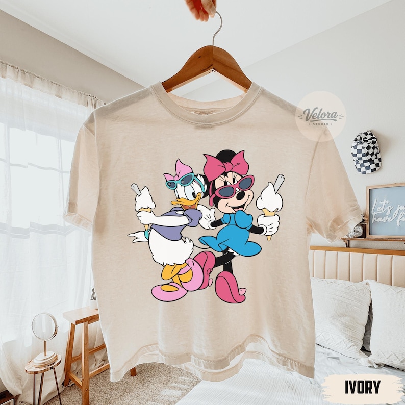 May include: Ivory-colored t-shirt featuring a graphic of Minnie Mouse and Daisy Duck holding ice cream cones. Minnie Mouse wears a pink bow and sunglasses, while Daisy Duck wears a purple bow and sunglasses. The shirt is hanging on a wooden hanger.