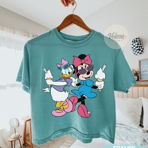 May include: Chalky mint green t-shirt featuring a cartoon illustration of Daisy Duck and Minnie Mouse wearing sunglasses and holding ice cream cones. The shirt is hanging on a wooden hanger.