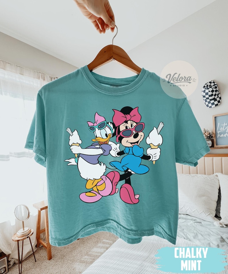 May include: Chalky mint green t-shirt featuring a cartoon illustration of Daisy Duck and Minnie Mouse wearing sunglasses and holding ice cream cones. The shirt is hanging on a wooden hanger.