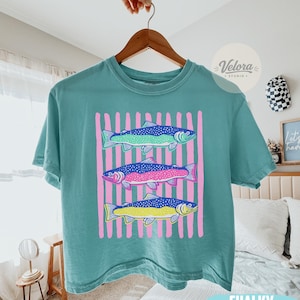 May include: A teal-colored t-shirt featuring a graphic of three colorful fish with pink vertical stripes in the background. The fish are in shades of green, blue, pink, and yellow. The shirt is hanging on a wooden hanger.