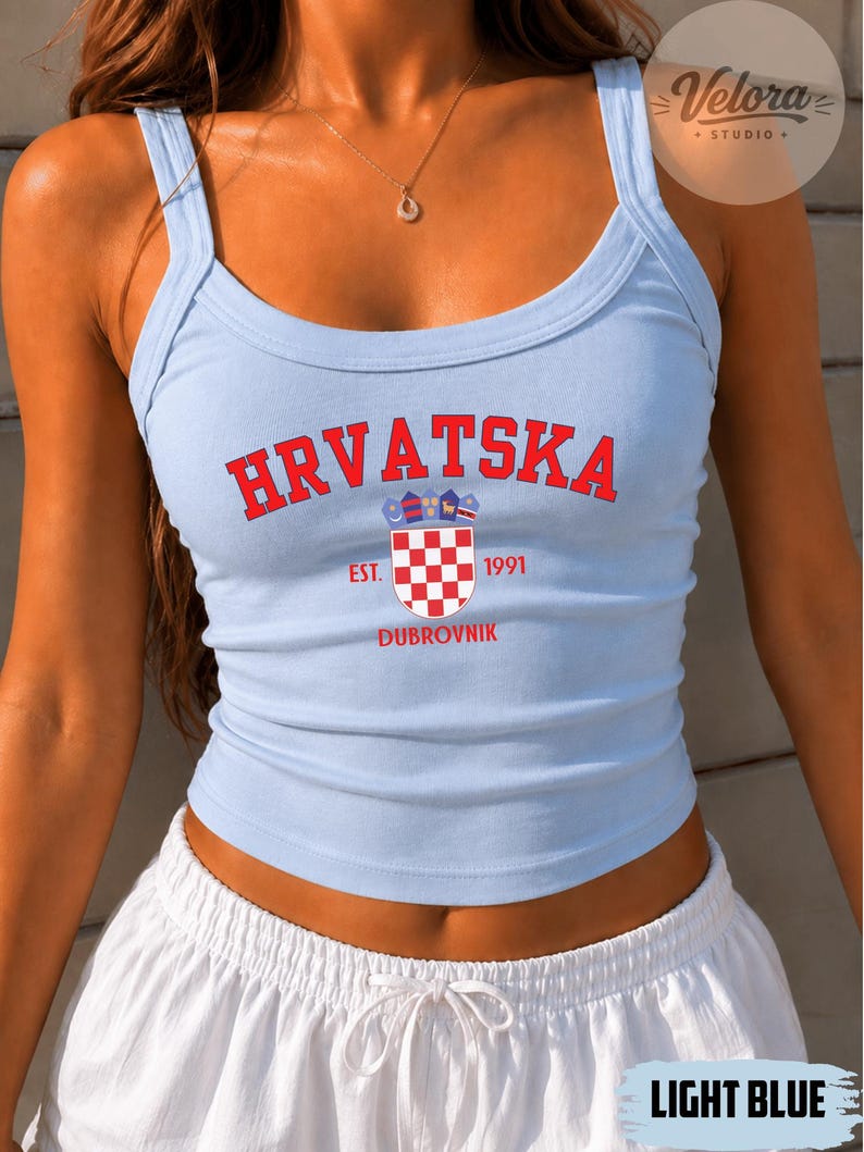 May include: A light blue tank top featuring the red text HRVATSKA in an arched font, a red and white checkered shield emblem, the text EST. 1991, and the word DUBROVNIK in red. The garment has a scoop neckline and thick shoulder straps.