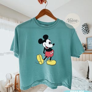 May include: A Chalky Mint colored t-shirt featuring a graphic of Mickey Mouse in his classic pose. Mickey is wearing red shorts, yellow shoes, and black gloves. The shirt is hanging on a wooden hanger.