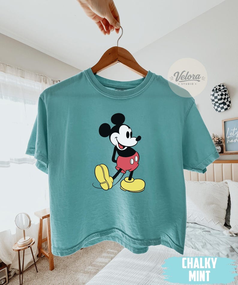 May include: A Chalky Mint colored t-shirt featuring a graphic of Mickey Mouse in his classic pose. Mickey is wearing red shorts, yellow shoes, and black gloves. The shirt is hanging on a wooden hanger.