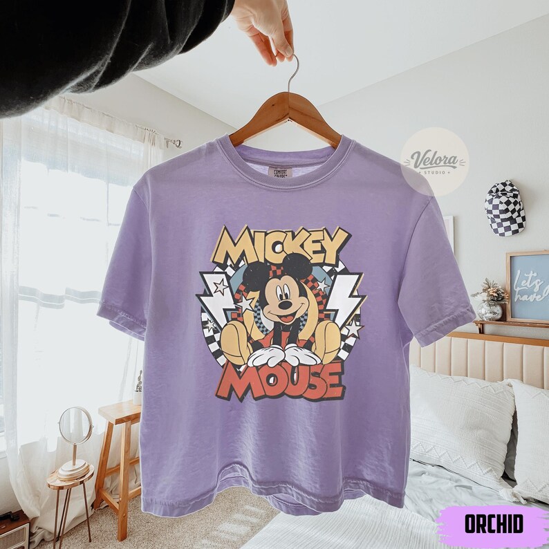 Puede incluir: Camiseta color orqu&iacute;dea con un gr&aacute;fico de Mickey Mouse. El dise&ntilde;o incluye las palabras "Mickey Mouse" en una fuente retro, con rayos y un fondo a cuadros. La camiseta est&aacute; colgada en una percha de madera.