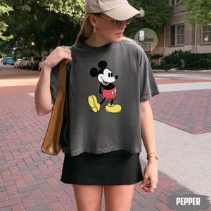 May include: A dark gray t-shirt featuring a classic Mickey Mouse graphic. The cartoon character is depicted in his iconic red shorts, yellow shoes, and white gloves. The word "PEPPER" is printed in white at the bottom right.