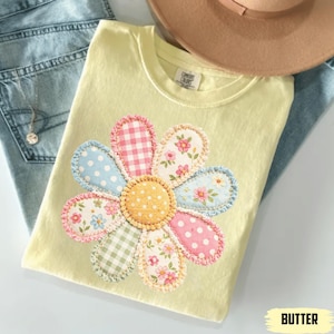 以下が含まれることがあります： 大きなアップリケの花柄が施された、明るい黄色のTシャツ。花びらは、水玉模様、ギンガムチェック、花柄など、さまざまな模様が特徴です。Tシャツは青いジーンズの上に折りたたまれており、背景には茶色の帽子が見えます。「BUTTER」の文字が右下にあります。