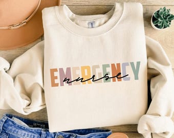 May include: Cream-colored sweatshirt with the words "EMERGENCY nurse" printed across the front in a colorful, block letter design. The word "nurse" is in a cursive font. The sweatshirt is folded on top of blue jeans and a brown hat.