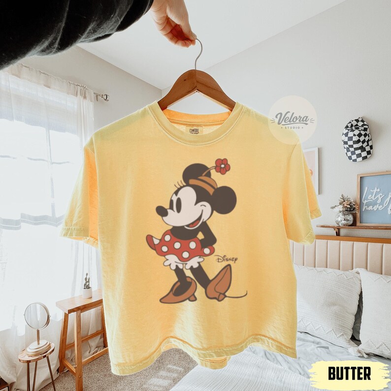 May include: Yellow t-shirt featuring a Minnie Mouse graphic. The graphic shows Minnie Mouse in a red polka dot skirt and brown shoes. The shirt has the word "Disney" printed below the graphic. The shirt is hanging on a wooden hanger.