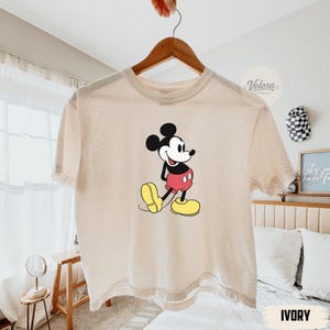 May include: Ivory-colored t-shirt featuring a classic Mickey Mouse graphic. The cartoon character is depicted in his iconic pose, wearing red shorts, yellow shoes, and black gloves. The shirt is hanging on a wooden hanger.