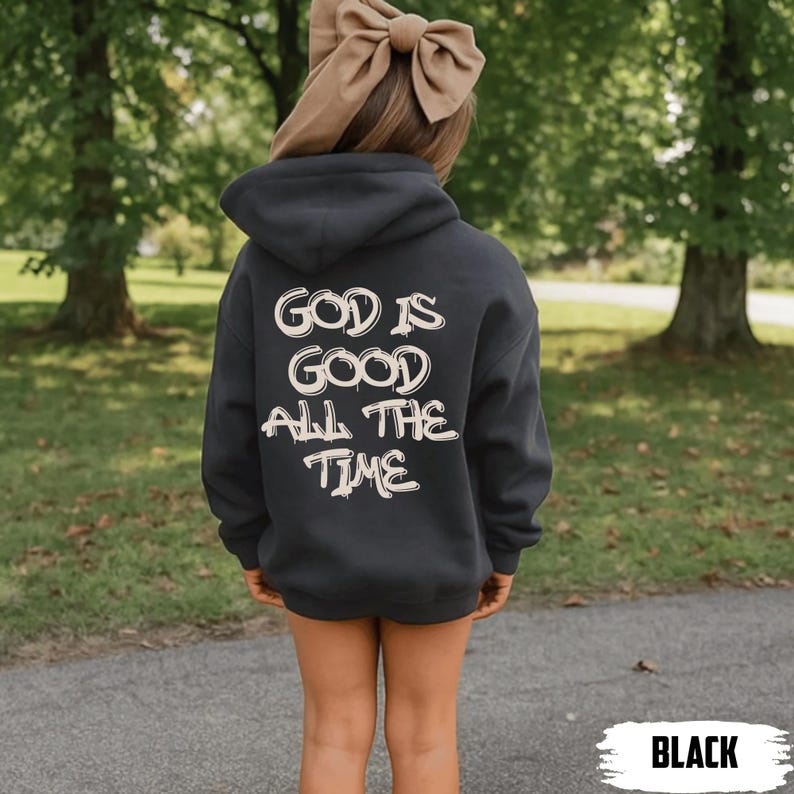 May include: A black hooded sweatshirt with the text "GOD IS GOOD ALL THE TIME" in a white, stylized font. The sweatshirt is worn by a person with a large tan bow in their hair. The word "BLACK" is in a white box at the bottom.