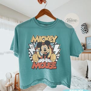 Puede incluir: Camiseta de color turquesa con un dise&ntilde;o de Mickey Mouse. El dise&ntilde;o incluye las palabras "Mickey Mouse" en un estilo retro, con rayos y estrellas. La camiseta est&aacute; colgada en una percha de madera.