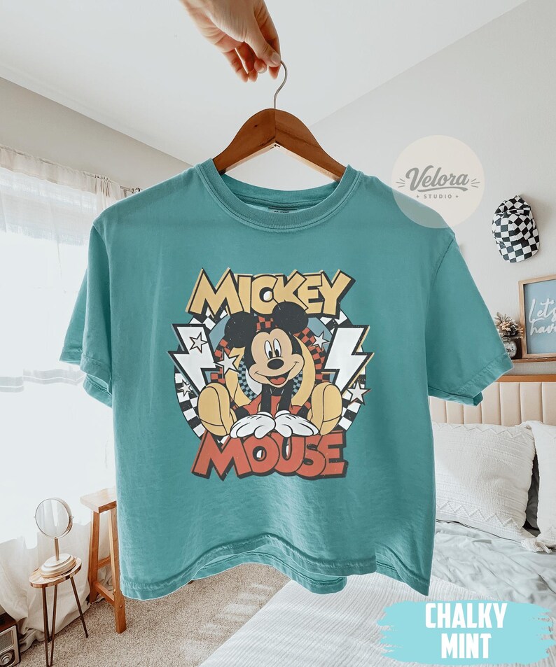 Puede incluir: Camiseta de color turquesa con un dise&ntilde;o de Mickey Mouse. El dise&ntilde;o incluye las palabras "Mickey Mouse" en un estilo retro, con rayos y estrellas. La camiseta est&aacute; colgada en una percha de madera.