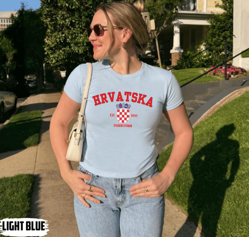 May include: Light blue t-shirt with the word "HRVATSKA" in red arched across the chest. Below the word is a Croatian coat of arms and the word "DUBROVNIK". The shirt is short-sleeved and made of a soft material.