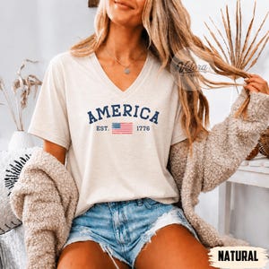 May include: A cream-colored V-neck t-shirt with the word "AMERICA" in navy blue, along with "EST. 1776" and a small American flag graphic. The shirt is paired with denim shorts and a beige cardigan.