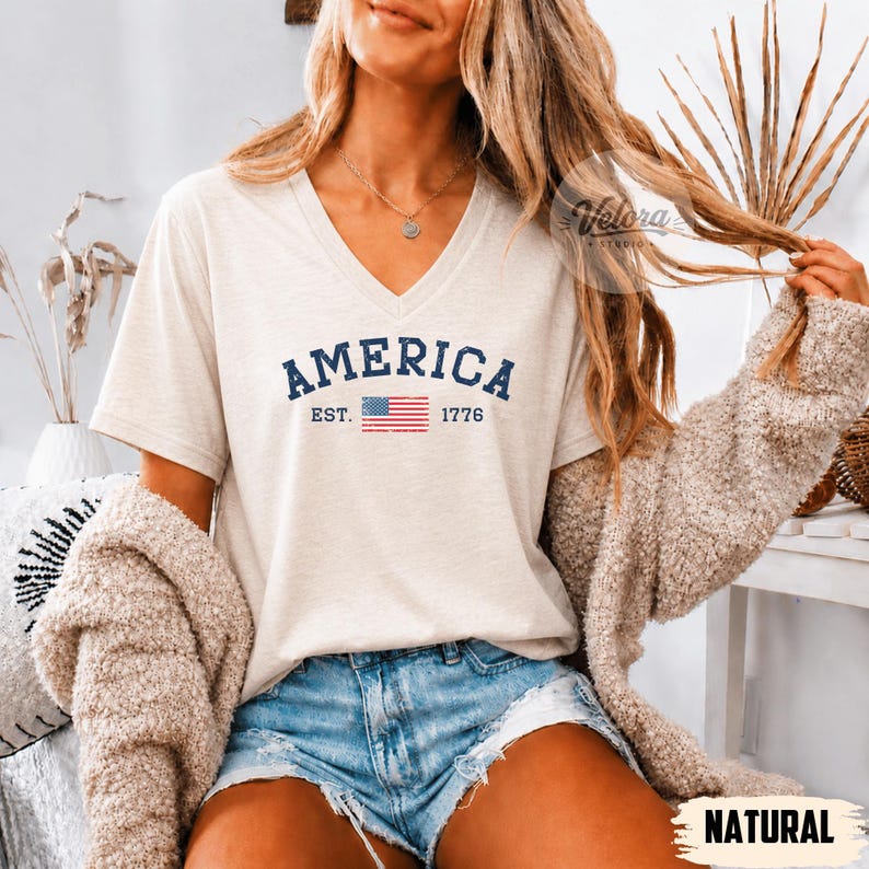 May include: A cream-colored V-neck t-shirt with the word "AMERICA" in navy blue, along with "EST. 1776" and a small American flag graphic. The shirt is paired with denim shorts and a beige cardigan.