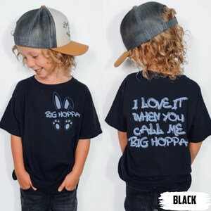 May include: Black t-shirt with the text "BIG HOPPA" and bunny ears and paw prints graphic. The back of the shirt says "I LOVE IT WHEN YOU CALL ME BIG HOPPA". The child is wearing a trucker hat with a tan brim.