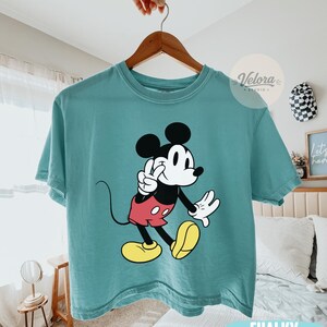 May include: A teal-colored t-shirt featuring a graphic of Mickey Mouse in a classic pose. Mickey is depicted with black ears, a white face, red shorts, and yellow shoes. The shirt has the words "Chalky Mint" printed at the bottom.
