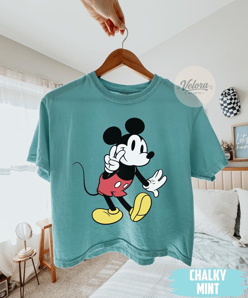 May include: A teal-colored t-shirt featuring a graphic of Mickey Mouse in a classic pose. Mickey is depicted with black ears, a white face, red shorts, and yellow shoes. The shirt has the words "Chalky Mint" printed at the bottom.