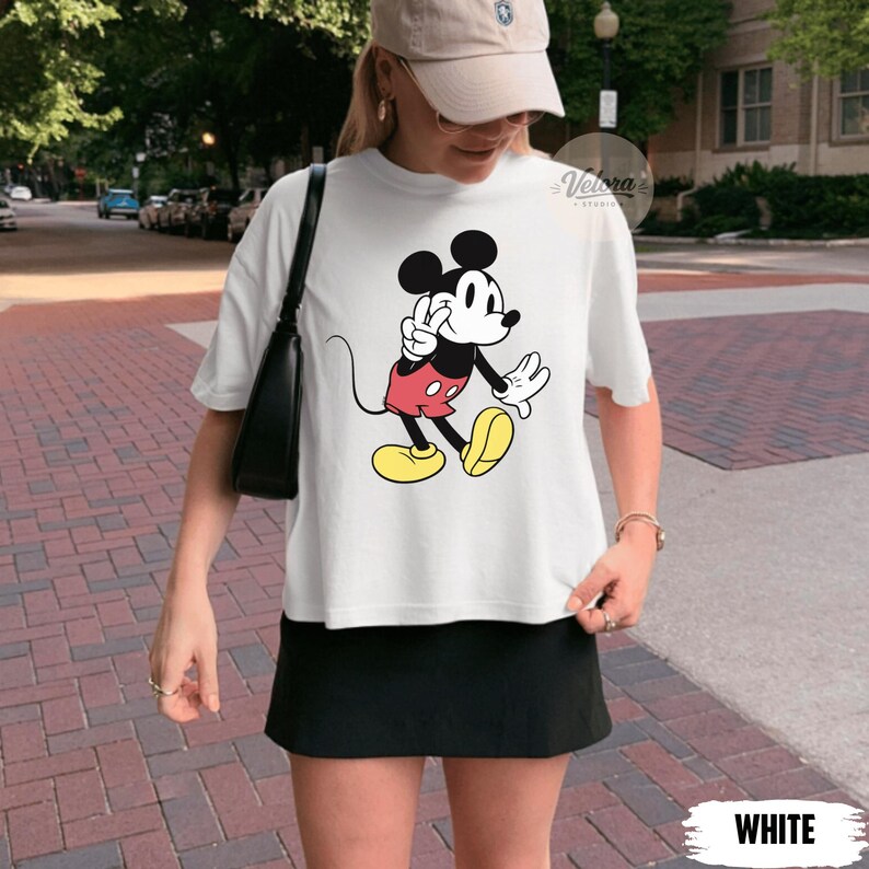 May include: White t-shirt featuring a classic Mickey Mouse graphic. The cartoon character is depicted in his iconic pose, wearing red shorts, yellow shoes, and white gloves. The shirt is paired with a black skirt.