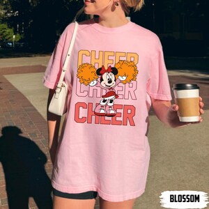 May include: Pale pink t-shirt with a cartoon Minnie Mouse design and the word "CHEER" in orange and red. Minnie Mouse is holding orange pom-poms. The shirt is a light pink colour.