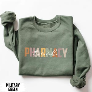 May include: A military green crewneck sweatshirt featuring the word PHARMACY in block letters with a muted, multi-colored palette. The word tech is written in a white cursive script layered over the center of the block letters.