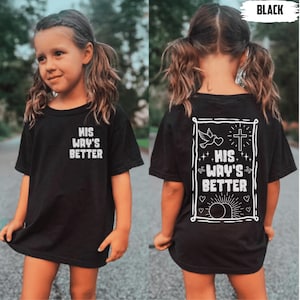 Può includere: T-shirt nera con la scritta bianca "HIS WAY'S BETTER" sul davanti e sul retro. Il disegno sul retro include una colomba, una croce e un'alba all'interno di una cornice bianca. La maglietta è indossata da una persona giovane.