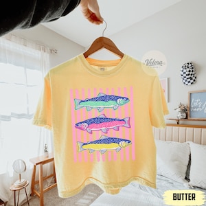 May include: Yellow t-shirt with a graphic of three colorful fish. The fish are blue, green, pink, and yellow, set against a pink striped background. The word "BUTTER" is at the bottom of the image.