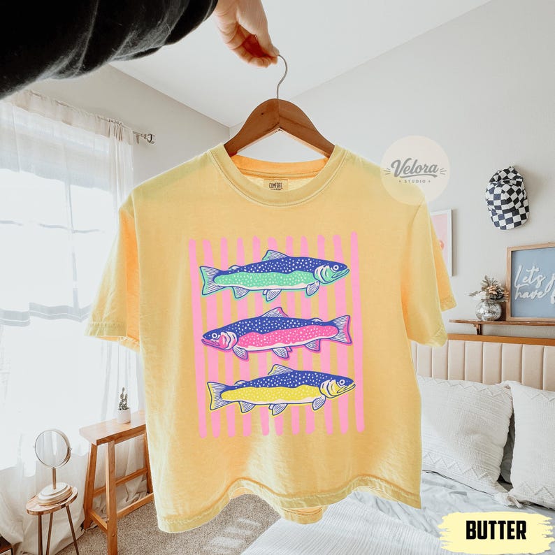 May include: Yellow t-shirt with a graphic of three colorful fish. The fish are blue, green, pink, and yellow, set against a pink striped background. The word "BUTTER" is at the bottom of the image.