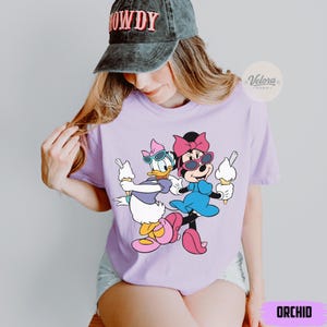 May include: A light purple t-shirt featuring a cartoon illustration of Minnie Mouse and Daisy Duck holding ice cream cones. Both characters are wearing sunglasses and bows. The word "ORCHID" is printed at the bottom of the shirt.