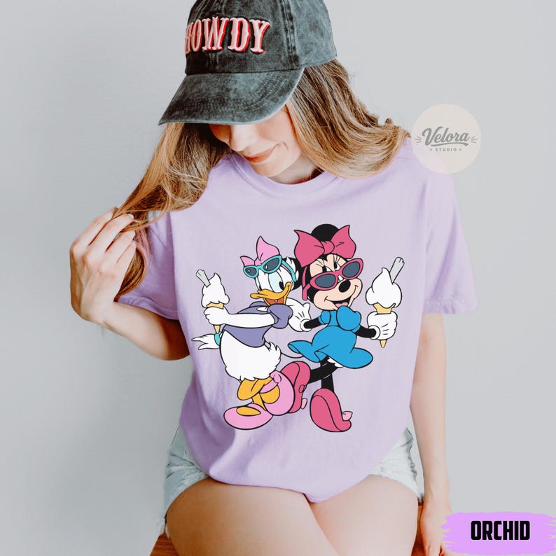 May include: A light purple t-shirt featuring a cartoon illustration of Minnie Mouse and Daisy Duck holding ice cream cones. Both characters are wearing sunglasses and bows. The word "ORCHID" is printed at the bottom of the shirt.