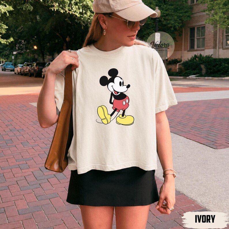 May include: Ivory-colored t-shirt featuring a classic Mickey Mouse graphic. The cartoon character is depicted in his iconic red shorts, yellow shoes, and black gloves. The shirt is paired with a black skirt.