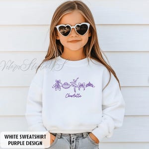 Puede incluir: Sudadera blanca con un dise&ntilde;o de ballet morado. El dise&ntilde;o incluye zapatillas de ballet, un tut&uacute;, una bailarina, lazos y una corona. El nombre "Charlotte" est&aacute; escrito en morado debajo. El texto "WHITE SWEATSHIRT PURPLE DESIGN" est&aacute; en la parte inferior.