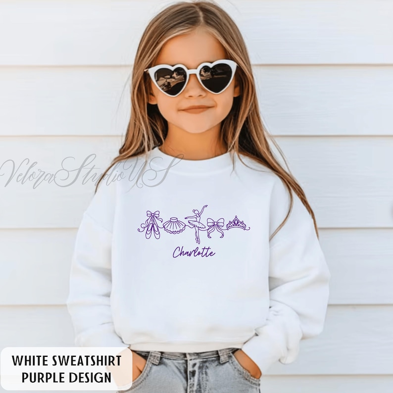 Puede incluir: Sudadera blanca con un dise&ntilde;o de ballet morado. El dise&ntilde;o incluye zapatillas de ballet, un tut&uacute;, una bailarina, lazos y una corona. El nombre "Charlotte" est&aacute; escrito en morado debajo. El texto "WHITE SWEATSHIRT PURPLE DESIGN" est&aacute; en la parte inferior.