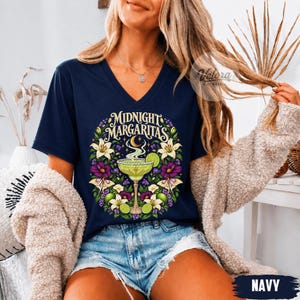 May include: A navy blue V-neck t-shirt featuring a graphic print of a margarita glass surrounded by white lilies, purple flowers, and moths. The text "MIDNIGHT MARGARITAS" is arched above the glass, which is garnished with a lime slice.