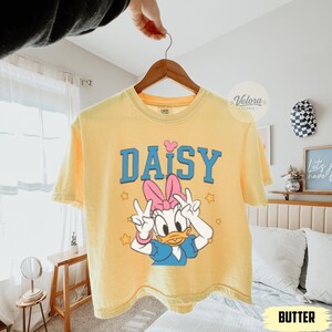 May include: Pale yellow t-shirt with a Daisy Duck graphic. The design features the word "DAISY" in blue, a pink bow, and Daisy Duck giving peace signs. The shirt is suspended from a wooden hanger.