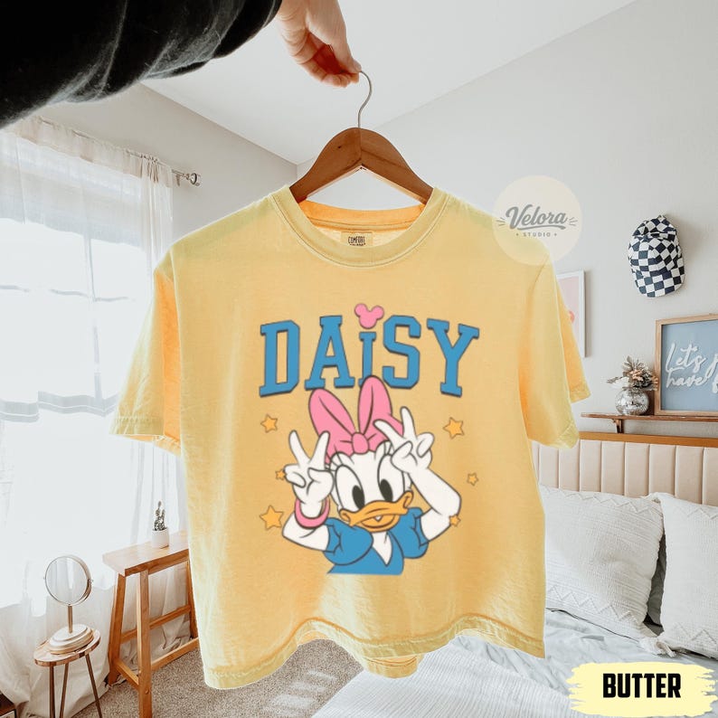 May include: Pale yellow t-shirt with a Daisy Duck graphic. The design features the word "DAISY" in blue, a pink bow, and Daisy Duck giving peace signs. The shirt is suspended from a wooden hanger.